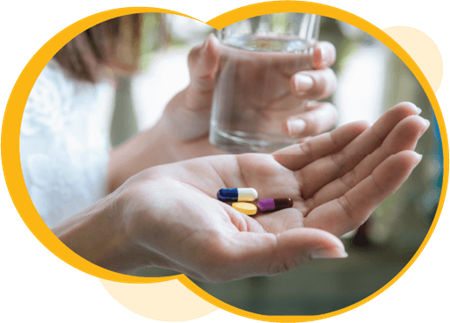 Gros plan d’une personne à la peau claire tenant un verre d’eau dans sa main gauche et trois pilules différentes dans sa main droite.